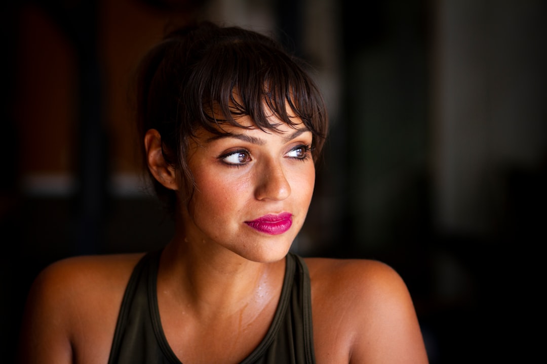 Woman with short dark hair and bangs looking to the side, wearing a sleeveless top, in a dimly lit indoor setting—her flawless look perfectly highlights mature skin makeup techniques.