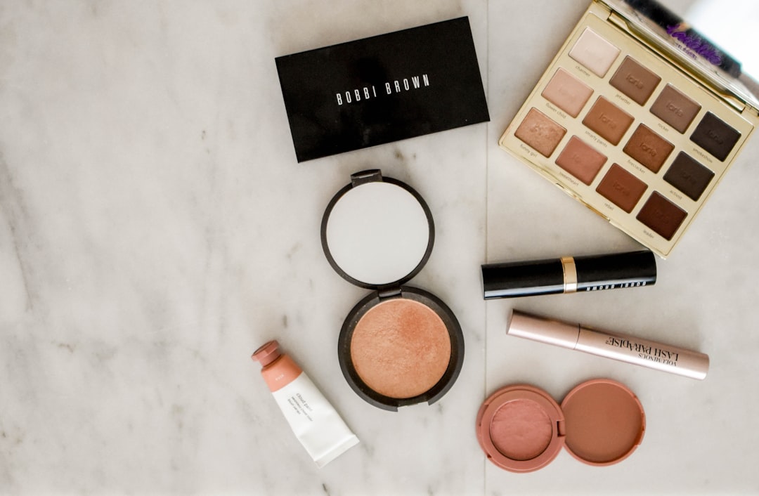 Various makeup products, including an eyeshadow palette, compact powder, blush, lipstick, cream tube, and mascara, are arranged on a light marble surface—perfect essentials for trying new makeup tips or mastering your highlight and contour routine.