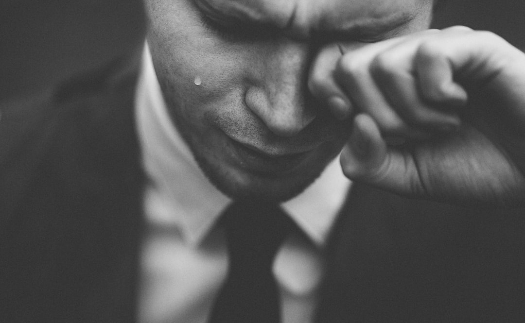 A man in a suit wipes his eye with his hand, a single tear on his cheek. The black and white close-up highlights not only emotion but also the refinement that comes from using the best men's grooming products.