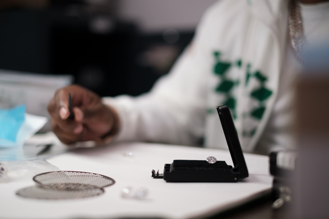 A person examines a loose diamond with tweezers next to a stunning piece of Messika diamond jewelry in an open box on a desk.