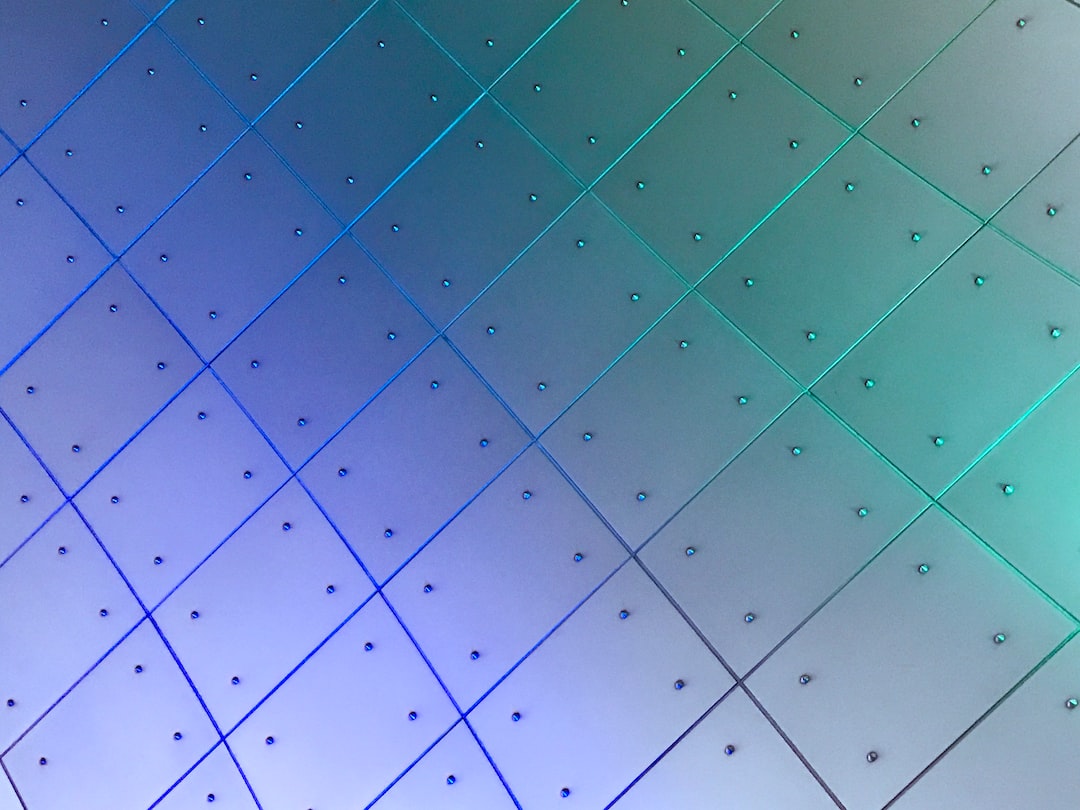 Close-up of a contemporary wall with metallic square panels arranged diagonally, each panel secured with visible bolts and illuminated with blue and green lighting reminiscent of Messika diamonds.