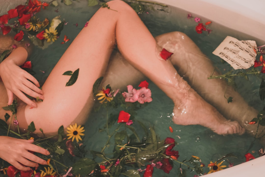 A person is lying in a bathtub filled with water, assorted flowers, and floating sheet music—an aesthetic scene that hints at relaxing Feuchtigkeitspflege for those seeking moisturizing tips. Only their legs and one hand are visible above the water.