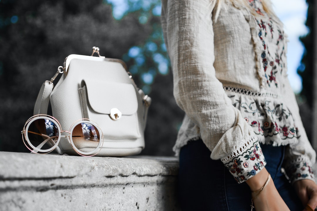 A woman in a floral embroidered blouse stands beside a white handbag and round sunglasses, showcasing fashion luxury on a stone surface outdoors.