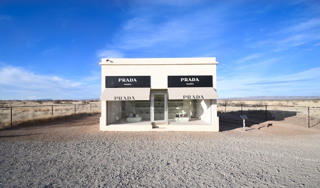 A small, standalone Prada store with large windows and black awnings stands isolated in a desert landscape under a blue sky, showcasing luxury handbags from one of the world’s most iconic expensive handbag brands.