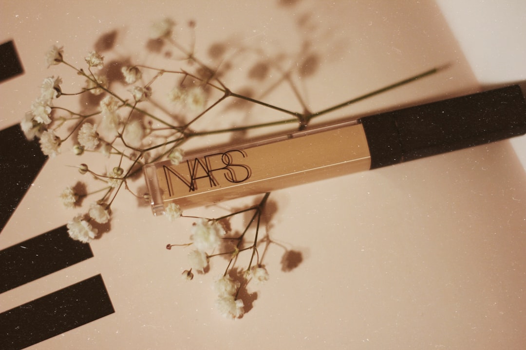 A NARS liquid concealer tube placed on a light surface with small white flowers arranged around it, offering flawless inspiration admired by beauty influencers.