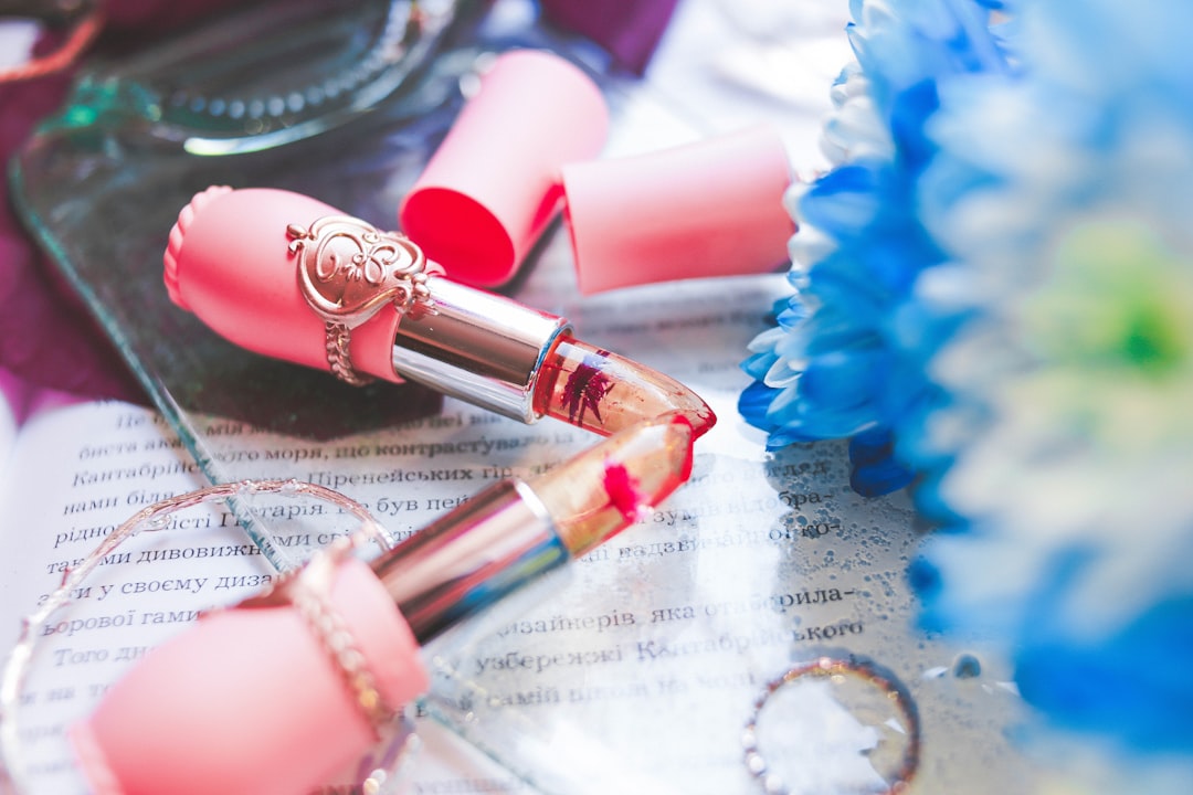 A broken pink lipstick, one of the must-have beauty products, lies on an open book, surrounded by a flower, a ring, and sunglasses.
