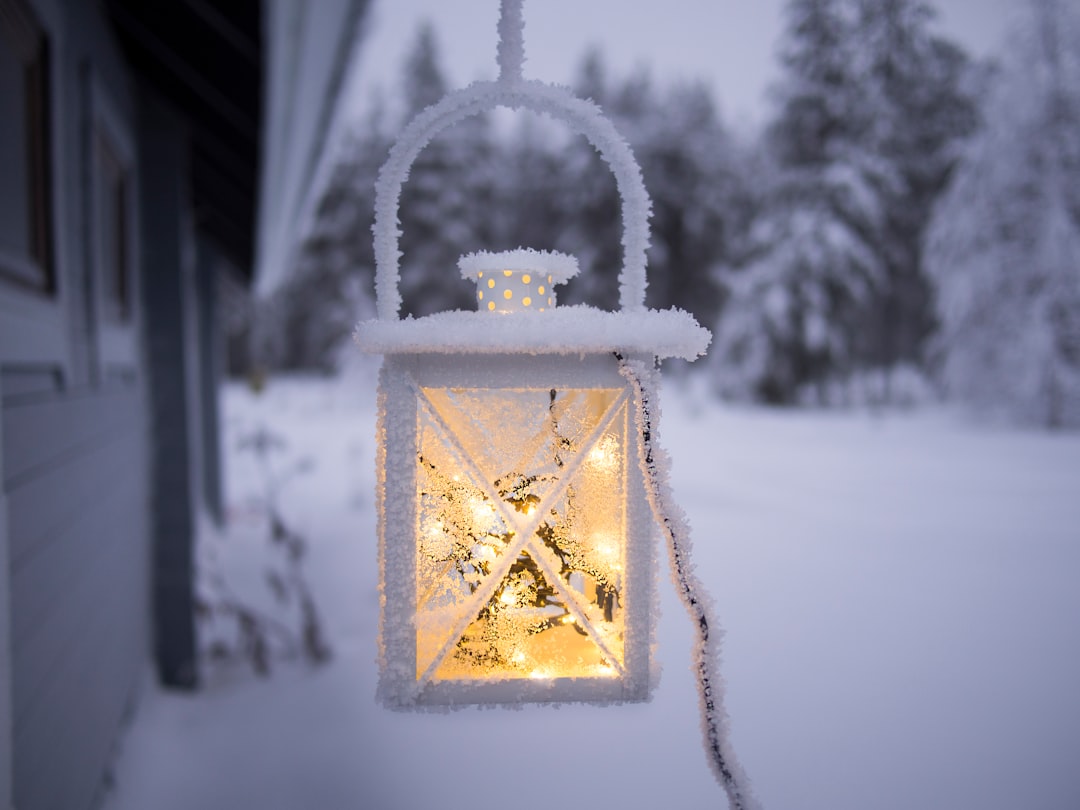 A frosty lantern with a glowing light hangs outside a building in a snowy, winter landscape with trees in the background, evoking the cozy comforts and glow of winter essentials.