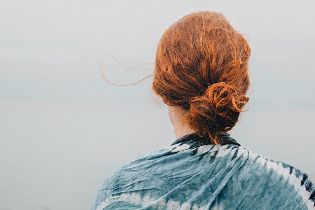 A person with red hair in a loose bun stands facing away, wrapped in a blue tie-dye shawl, looking out toward a misty, gray sky and water—embracing natural beauty without concern for unwanted hair.