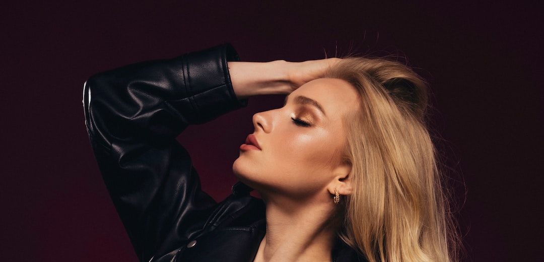 A woman with long blonde hair wearing a black leather jacket poses with her eyes closed and one hand in her hair, evoking feminine aesthetics against a dark background.