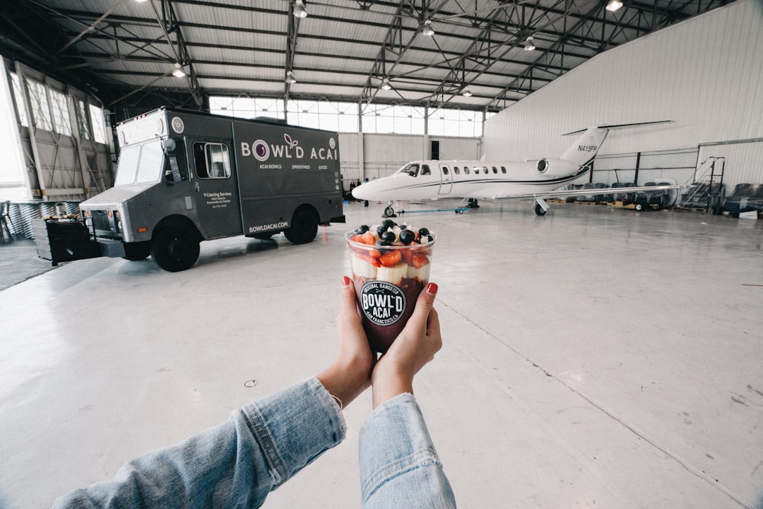 A person holds a bowl of acai topped with fruit in a hangar, embracing self-improvement, with a private jet and a food truck in the background.