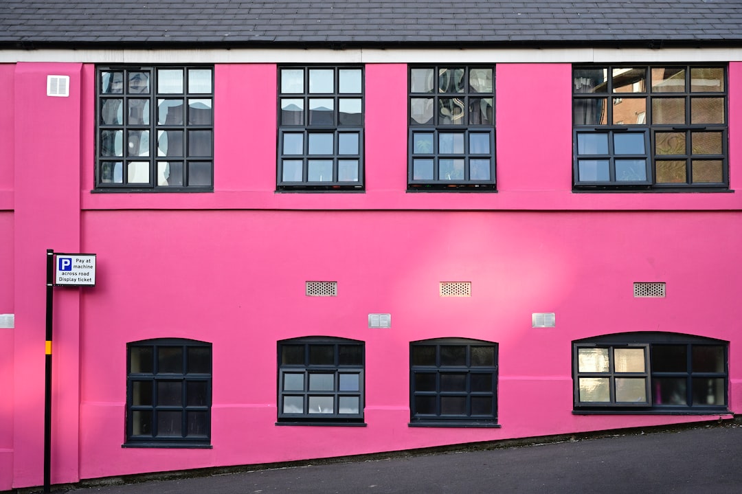 A building with bright pink exterior walls, reminiscent of Pomellato’s bold statement jewelry, features two rows of black-framed windows; a parking sign is mounted on the left side of the wall.