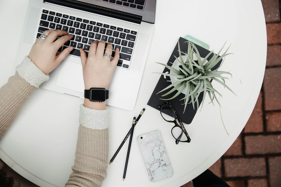 A person types on a laptop at a round white table with a plant, glasses, two pencils, a closed notebook, and a smartphone nearby—perfect for preserving time and focusing on tasks.
