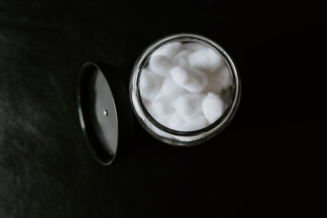 A glass jar filled with white cotton balls, perfect for make-up removers, sits on a dark surface next to its black lid.