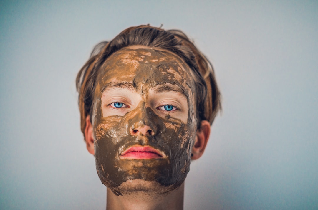 A person with light brown hair and blue eyes faces the camera, wearing a thick brown facial mud mask—one of many beauty products used to achieve radiant skin. The background is plain and neutral.