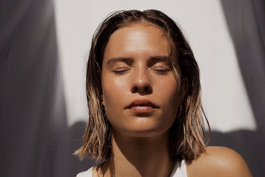 A woman with short, wet hair stands in sunlight with her eyes closed, her radiant skin glowing against a white background.