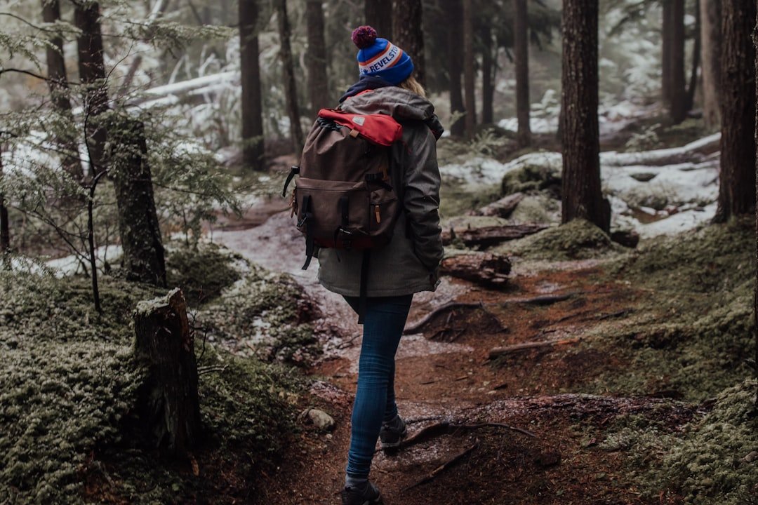 Person wearing a backpack and winter hat walks along a forest trail surrounded by trees and patches of snow, perhaps pondering tips and tricks to reduce facial swelling after outdoor adventures.