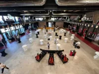 Spacious luxury indoor venue with scattered high tables, exhibits lining the walls, and two state-of-the-art racing simulators at the forefront; a few people are setting up.
