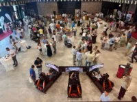 A large group of people socializes around tables at a luxury indoor event, with three car racing simulators in the foreground and food stations in the background.