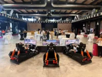 Three people use luxury racing simulators with large screens in a spacious event hall, while attendees gather and mingle near tables in the background.
