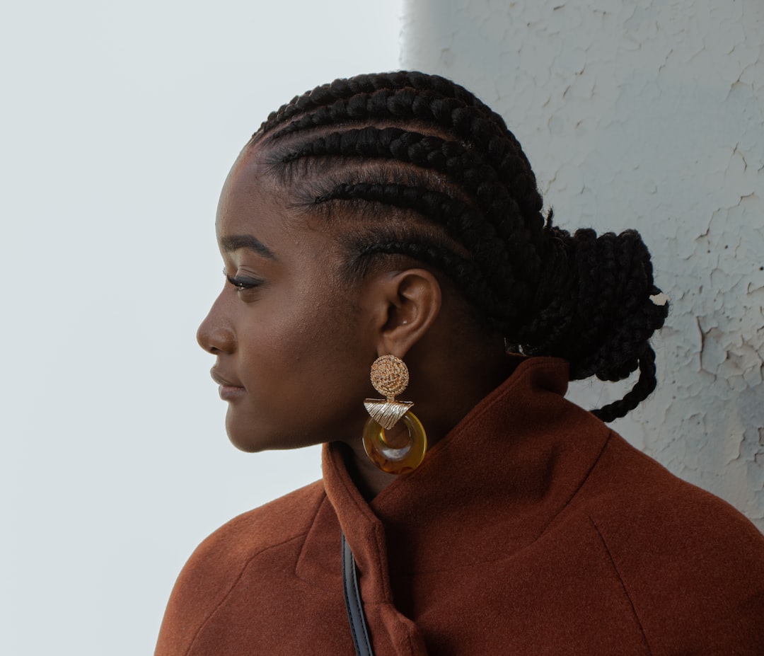 A woman with braided hair and large earrings exudes avant-garde fashion in a rust-colored coat, looking to the left before a textured, light-colored wall.
