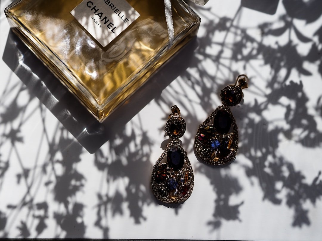A bottle of Chanel perfume and a pair of ornate, gemstone earrings—showcasing Italian craftsmanship—rest on a white surface with floral shadows, embodying modern elegance.
