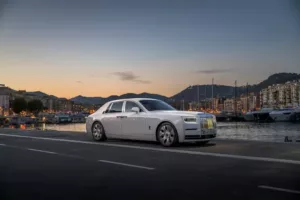 A luxury white Rolls-Royce sedan is parked by a marina at sunset, with yachts and waterfront buildings in the background.