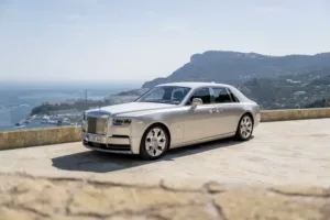 A silver luxury sedan is parked on a stone terrace overlooking a stunning coastline, with rolling hills and shimmering water in the background.