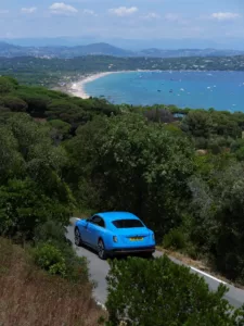 A blue luxury car glides along a narrow road surrounded by lush greenery, overlooking a coastal bay dotted with boats and a distant shoreline beneath the clear blue sky.
