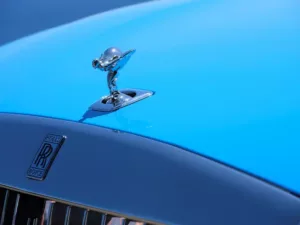 Close-up of the luxury Rolls-Royce hood ornament, the Spirit of Ecstasy, gleaming atop a bright blue car with the Rolls-Royce emblem visible below.