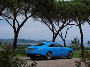 A blue luxury coupe car is parked on a dirt road under tall trees, offering a scenic view of sparkling water and rolling hills in the background.