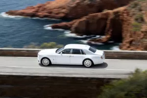 A white luxury sedan cruises along a scenic coastal road, with rocky cliffs and the sparkling ocean providing a breathtaking backdrop.