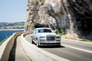 A luxury silver Rolls-Royce car glides along a coastal road beside a rocky cliff and the sea under a clear sky.