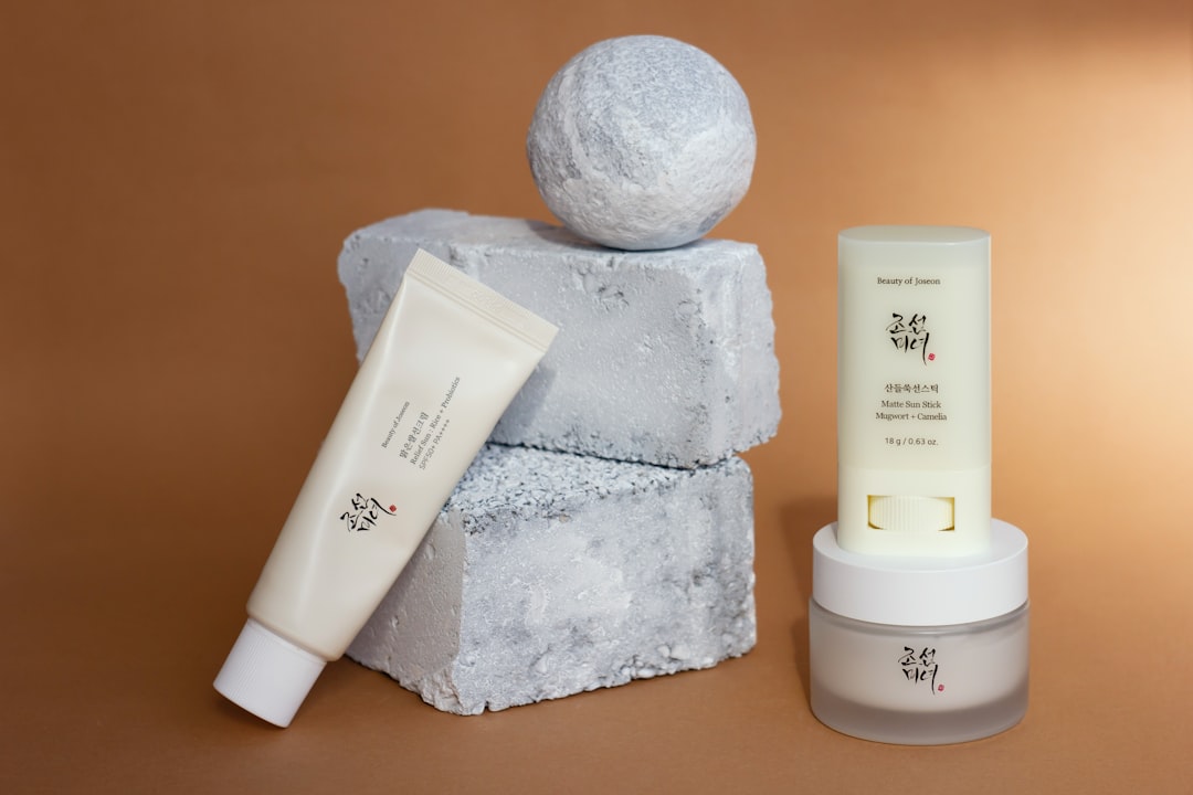 Three skincare products with Korean labels are displayed next to two stacked gray textured blocks and a gray spherical object, all set against a brown background—perfect for sun lovers seeking safe tanning solutions.