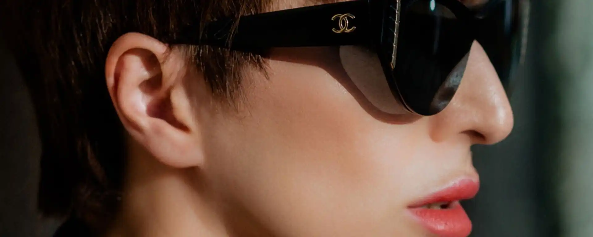 Close-up of a person's face wearing large black luxury Chanel sunglasses, with short brown hair and red lipstick visible.
