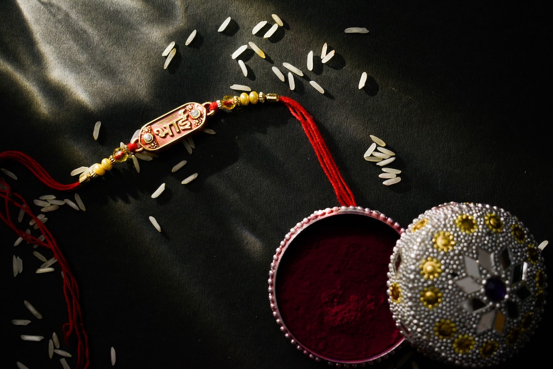 A decorative rakhi, scattered rice grains, and an ornate box containing red powder are arranged on a dark surface under streaks of light, making it a perfect choice for jewelry gifts or gifts for every occasion.