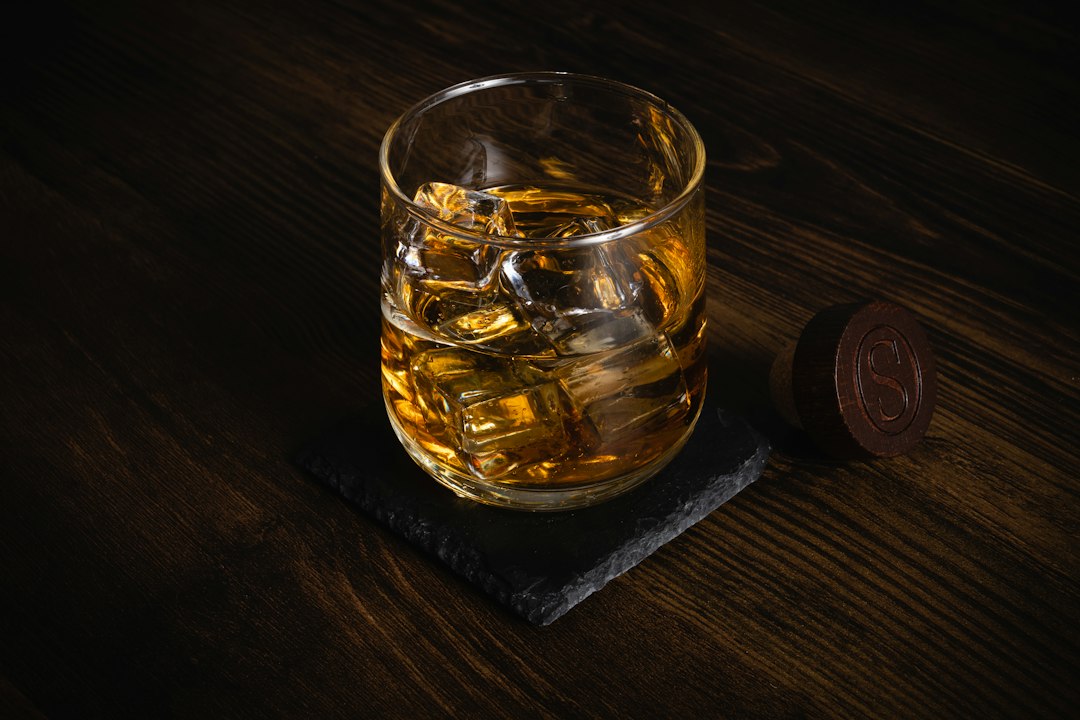 A glass of whiskey with ice cubes sits on a dark stone coaster, placed on a wooden surface next to a bottle cap—perfect for those who appreciate cocktail rings that shine bright and elevate your style.
