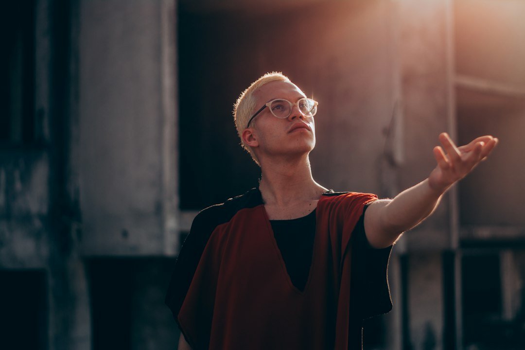 A person with short blond hair and glasses extends their arm outward while standing outdoors in dramatic sunlight, radiating ethical elegance as they shine bright.