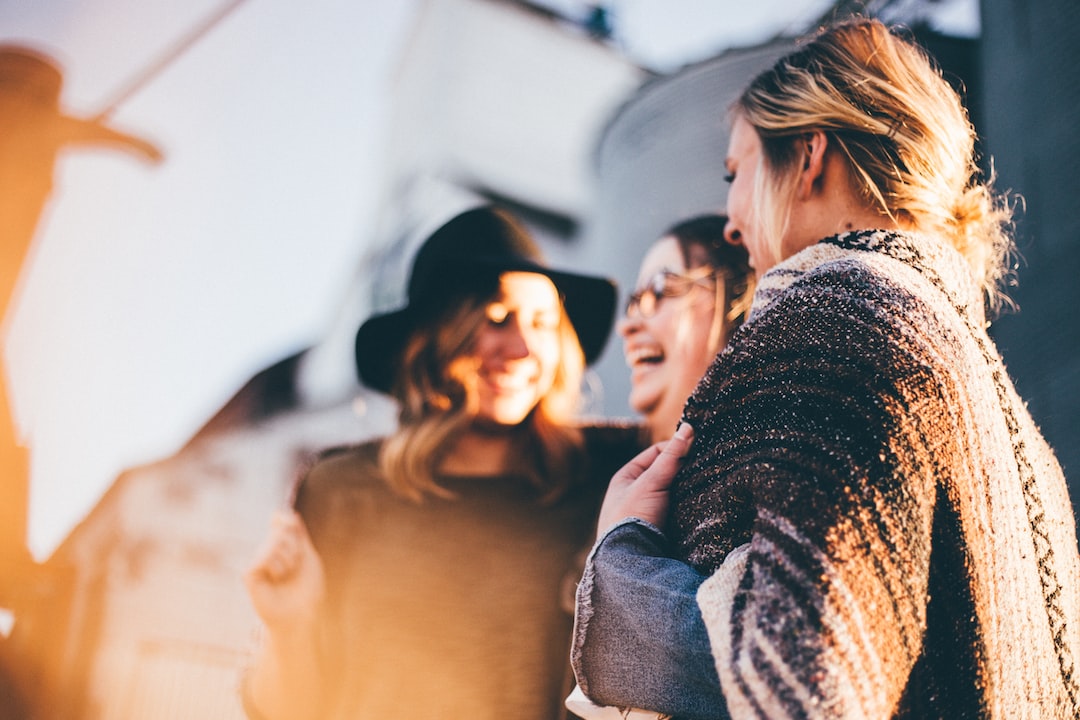 Three women stand outdoors, smiling and laughing together. One wears a black hat, another wears glasses, and all showcase the latest jewelry trends with casual, layered clothing.