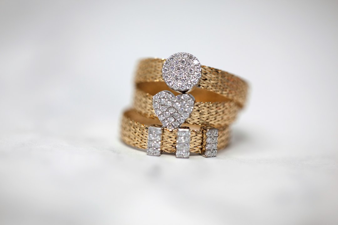 Three gold rings with textured bands and diamond-studded shapes—circle, heart, and rectangles—are stacked together on a white surface, showcasing the elegance of sustainable jewelry.