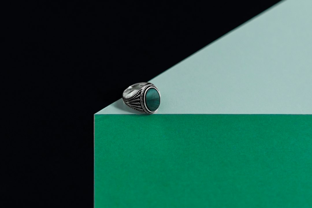 A silver ring with a green oval stone rests on green and mint-colored geometric surfaces, capturing the allure of the latest jewelry trends against a black background.