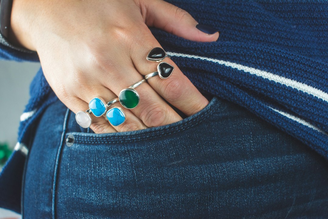 A hand adorned with the best jewelry—several colorful gemstone rings—rests on the pocket of blue jeans, while the person wears a blue sweater with white stripes.