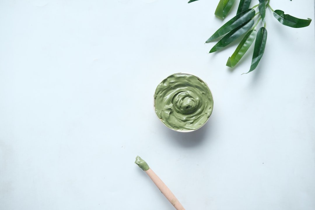 A bowl of green cream or paste sits on a white surface with a wooden spatula and green leaves nearby, perfect for skincare tips and pore reduction routines.