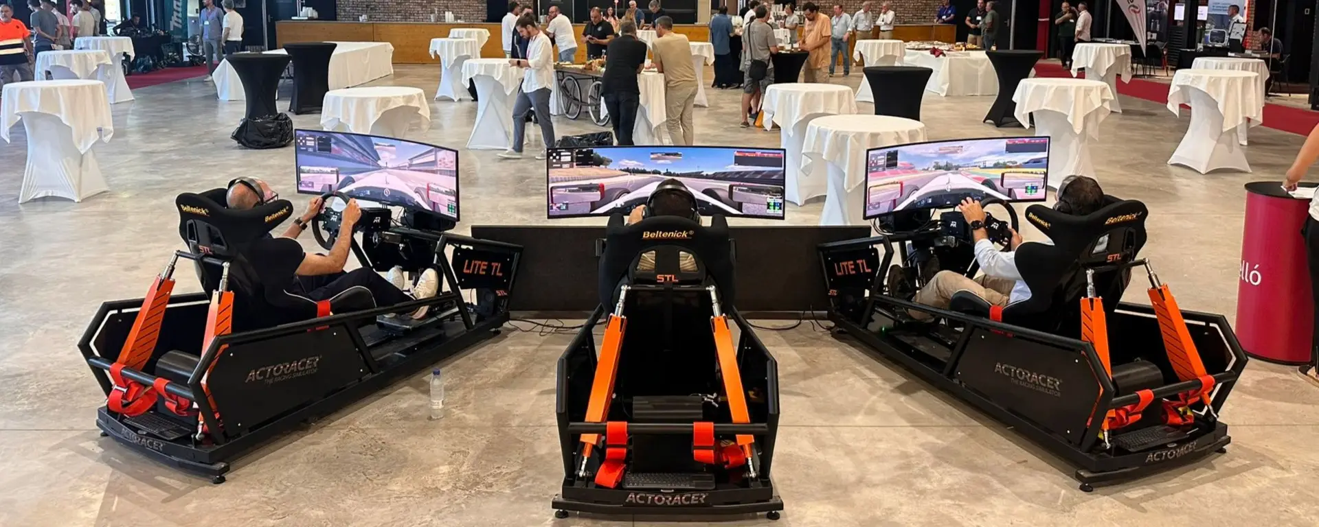 Three people sit in luxury racing simulator seats with steering wheels and screens, participating in a driving simulation event. Tables and people are visible in the background.