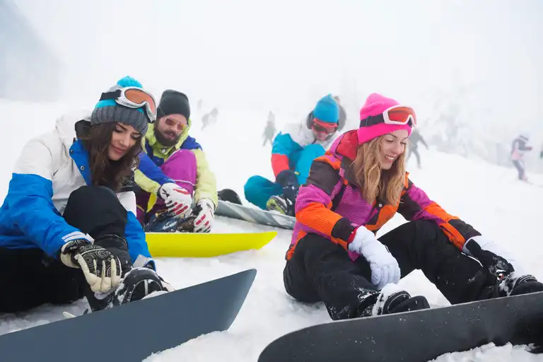 A group of people in colorful, luxury winter clothing and helmets sit in the snow, strapping on their snowboards.