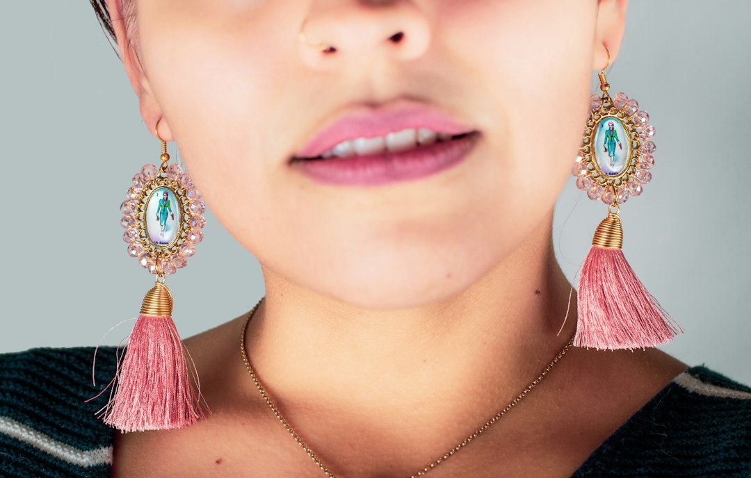 A close-up of a person wearing stunning earrings with pink tassels and religious medallions, plus a nose ring, showing only the lower half of the face.