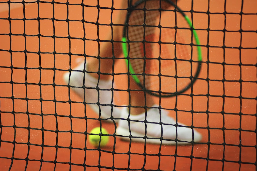 A tennis racket, player’s legs in white shoes, and a tennis ball are visible behind a black net on a clay tennis court, capturing the elegance of the game.