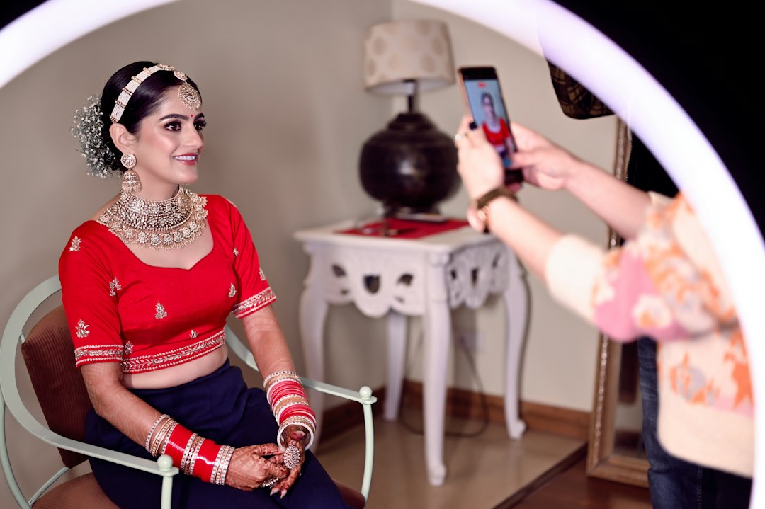 A bride in bridal jewelry and a red blouse sits on a chair, smiling as someone takes her photo with a smartphone under a ring light.
