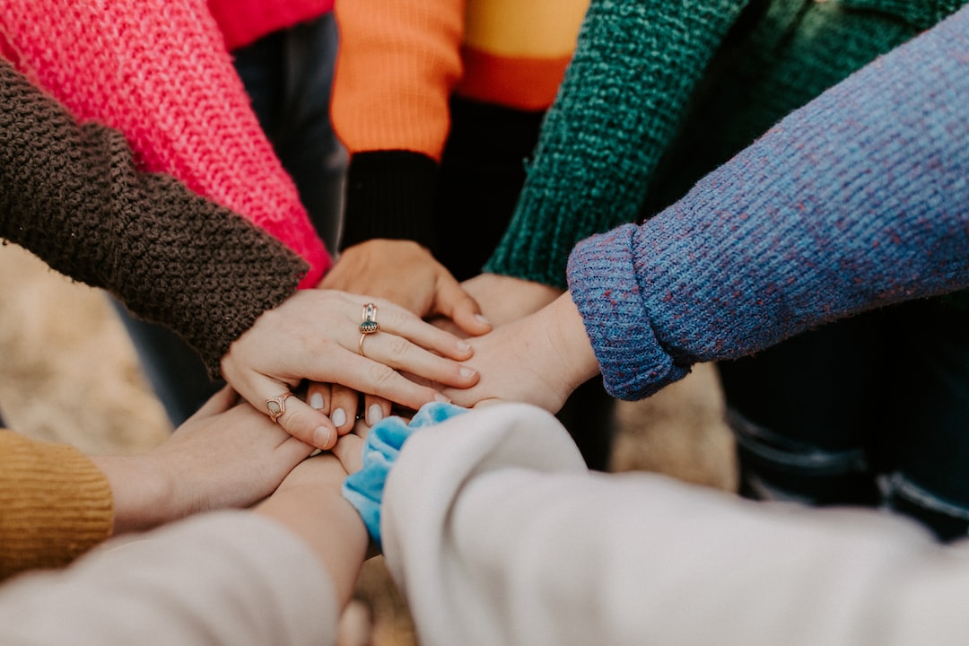 A group of people wearing colorful sweaters stand in a circle with their hands stacked together in the center, their rings gleaming as if after a recent jewelry cleaning.