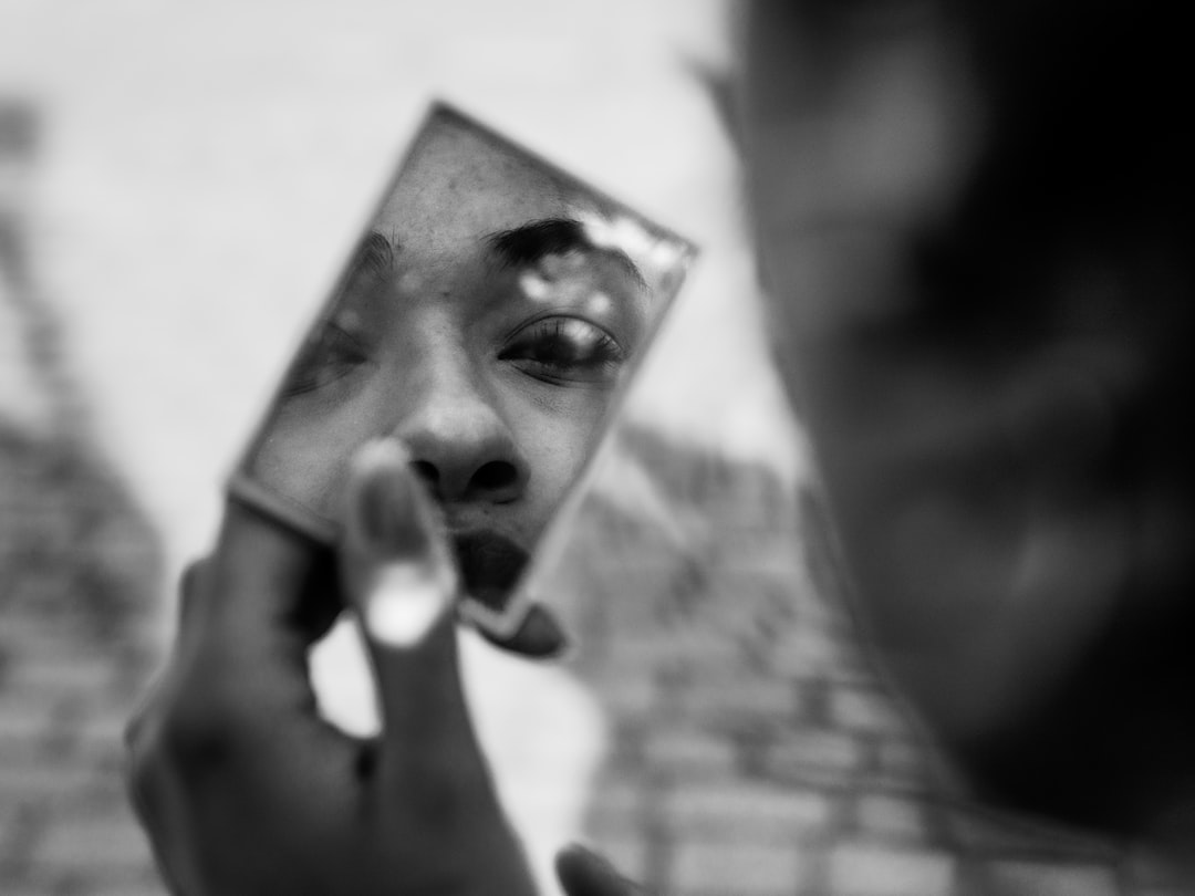 A person holds a small mirror, reflecting part of their face with a focus on one eye, capturing the edge and intrigue found in Stephen Webster's fine jewelry design. The image is in black and white.