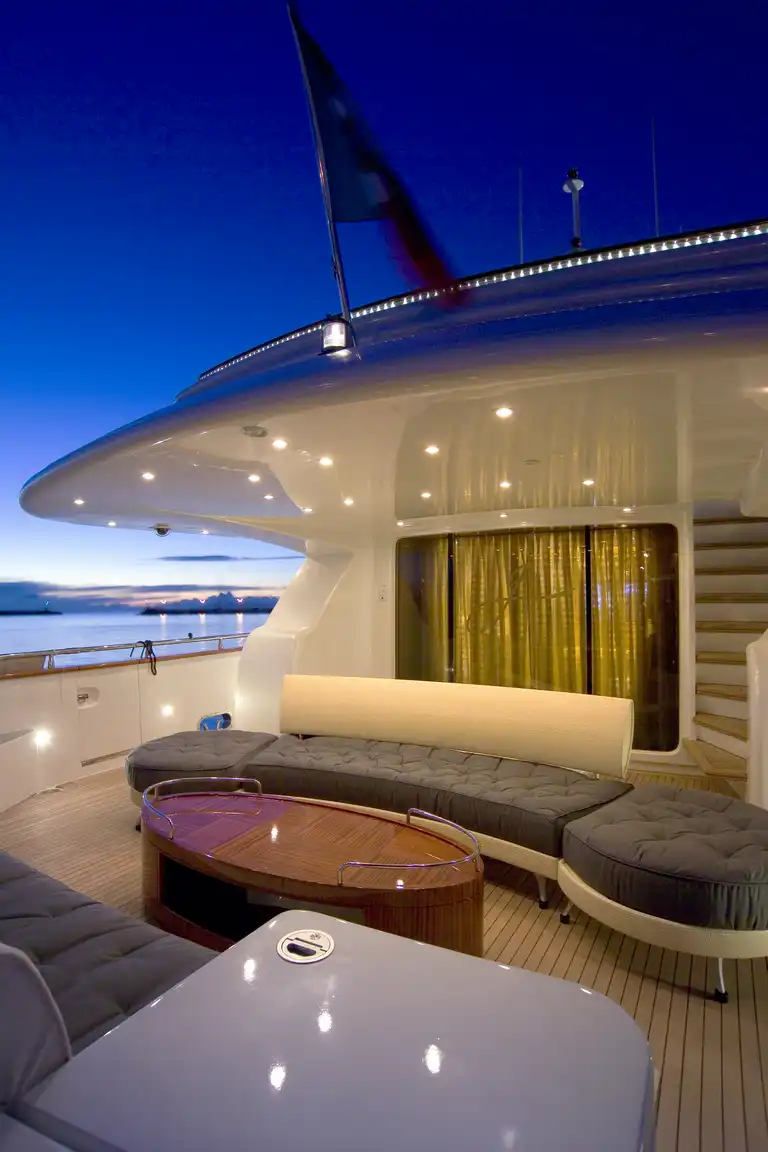 Modern luxury yacht deck at dusk with built-in seating, a wooden table, and a flag visible on the upper deck, overlooking calm water and a clear sky.